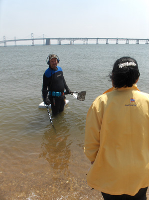 2 photos show Nanta still standing in the water.  A man is standing deeper in the water and talking with her.  He is wearing a 'wet suit' (covering his body for warmth) and the water is above his knees.  He wears headphones *to hear the metal detector) and has a metal detector with a hose, and a little floating raft to hold his treasure.